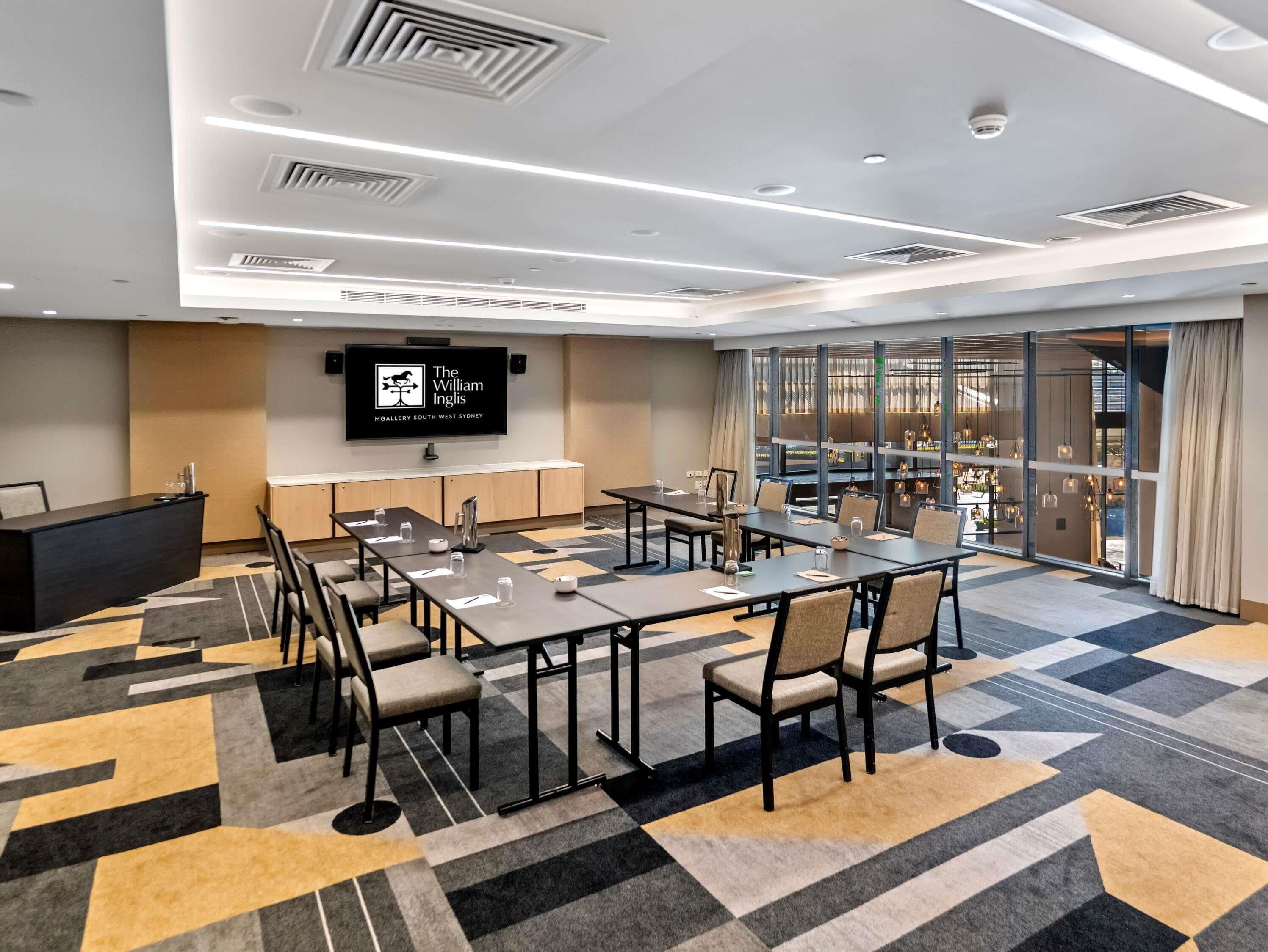 A conference room with a large screen displaying the hotel name, multiple tables with chairs, and a patterned carpet.