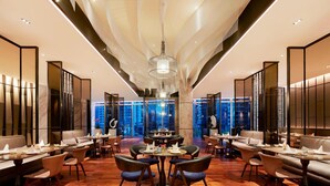 Un restaurant moderne avec des tables rondes et des sièges rembourrés, un plafond haut orné d’un luminaire original et une vue sur le paysage urbain grâce à de grandes fenêtres.