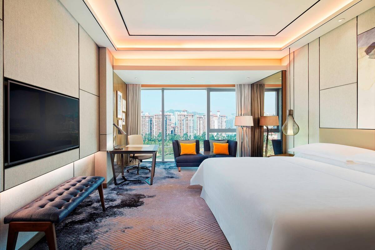 Ein Hotelzimmer mit einem großen Bett, einem Schreibtisch, einem Sessel, einer Couch mit orangenen Kissen, einem Fernseher und einem großen Fenster mit Blick auf die Stadt.