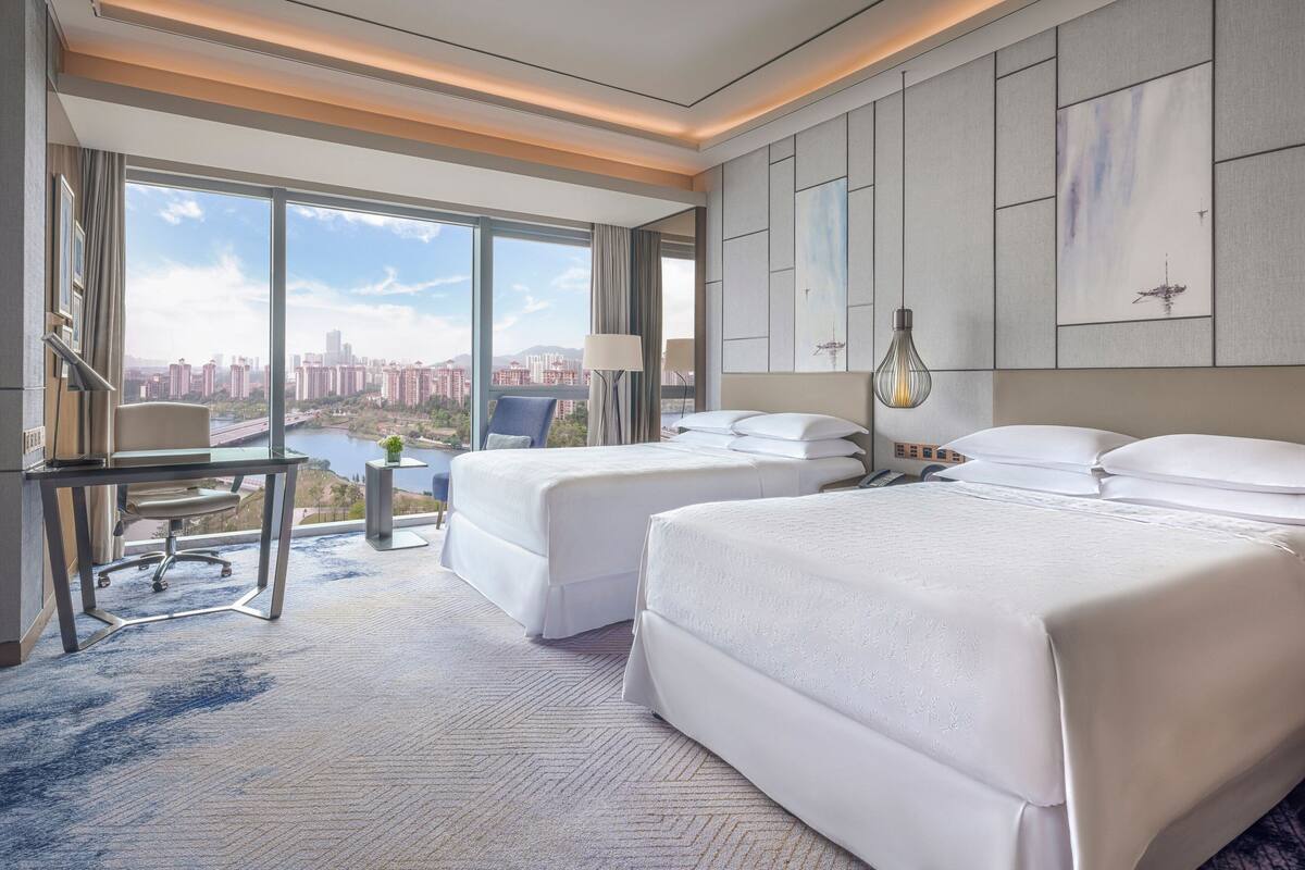 Ein Hotelzimmer mit zwei Betten, einem Schreibtisch und einem großen Fenster mit Blick auf die Stadt.