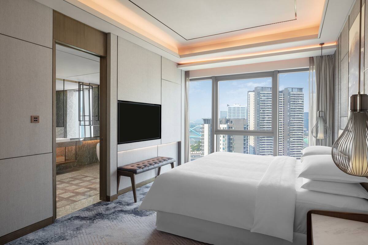 Ein modernes Hotelzimmer mit einem großen Bett, einem Flachbildfernseher, einer Sitzbank und Blick auf die Stadtlandschaft.