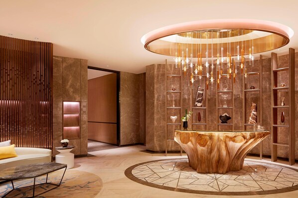 A lobby with a central wooden reception desk, a modern chandelier, and marble walls with shelves displaying decorative items.