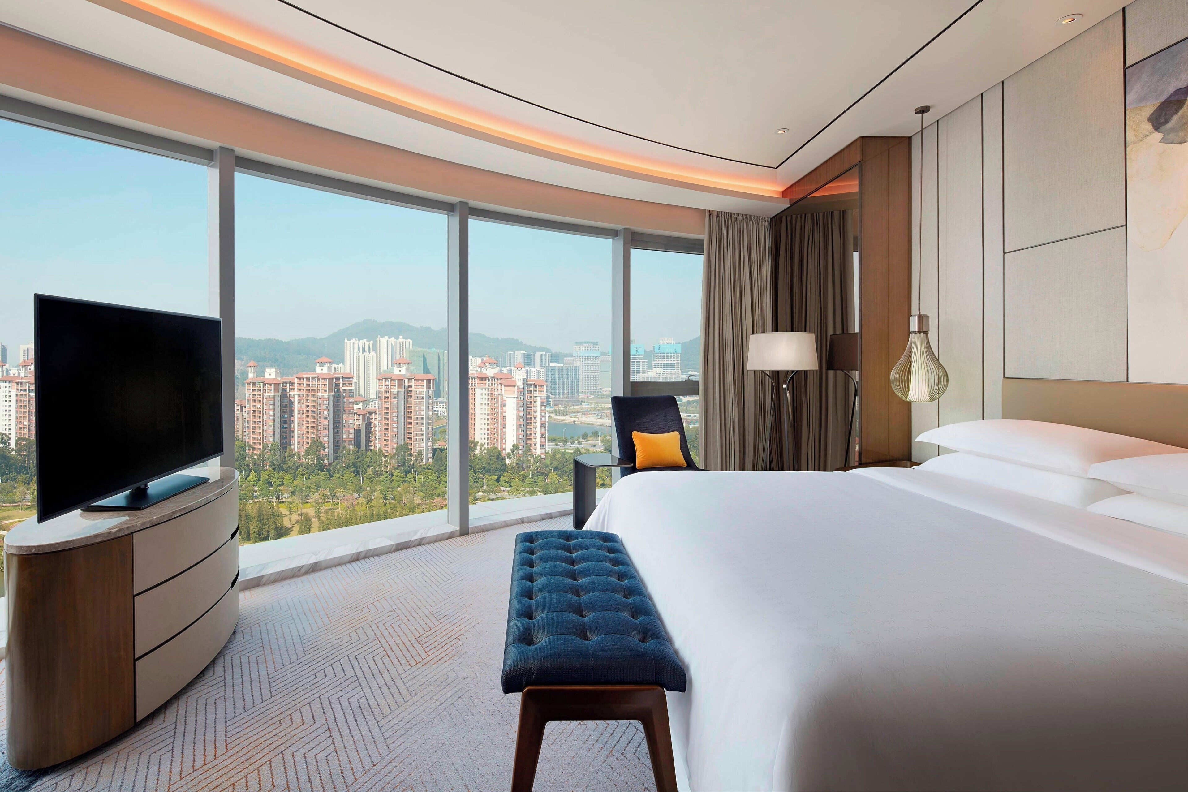 A modern hotel room with a large bed, a flat-screen TV, a bench, and a city view.