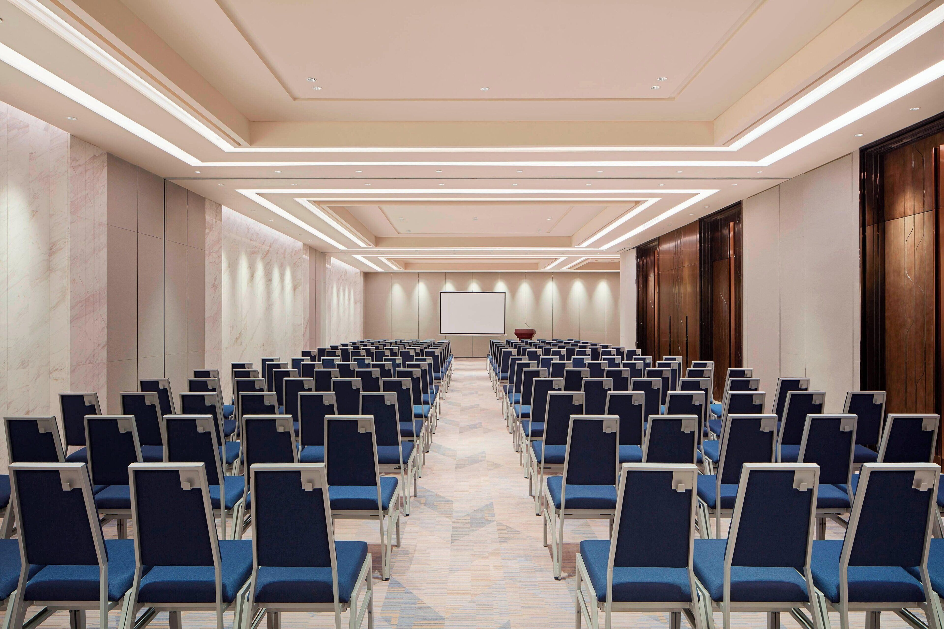 A conference room with rows of blue chairs, a large screen at the front, and a patterned carpet.