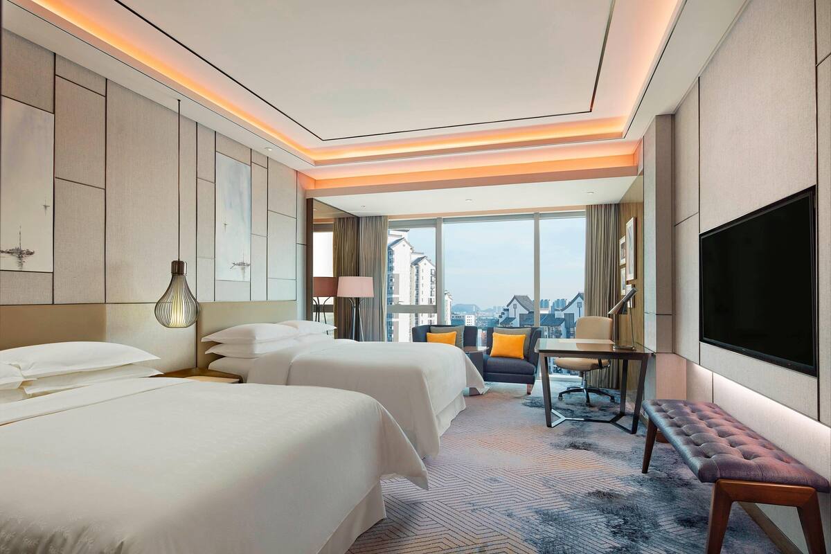 Ein Hotelzimmer mit zwei Betten, einem Schreibtisch, einer Bank, einem Fernseher und einem großen Fenster mit Blick auf die Stadt.