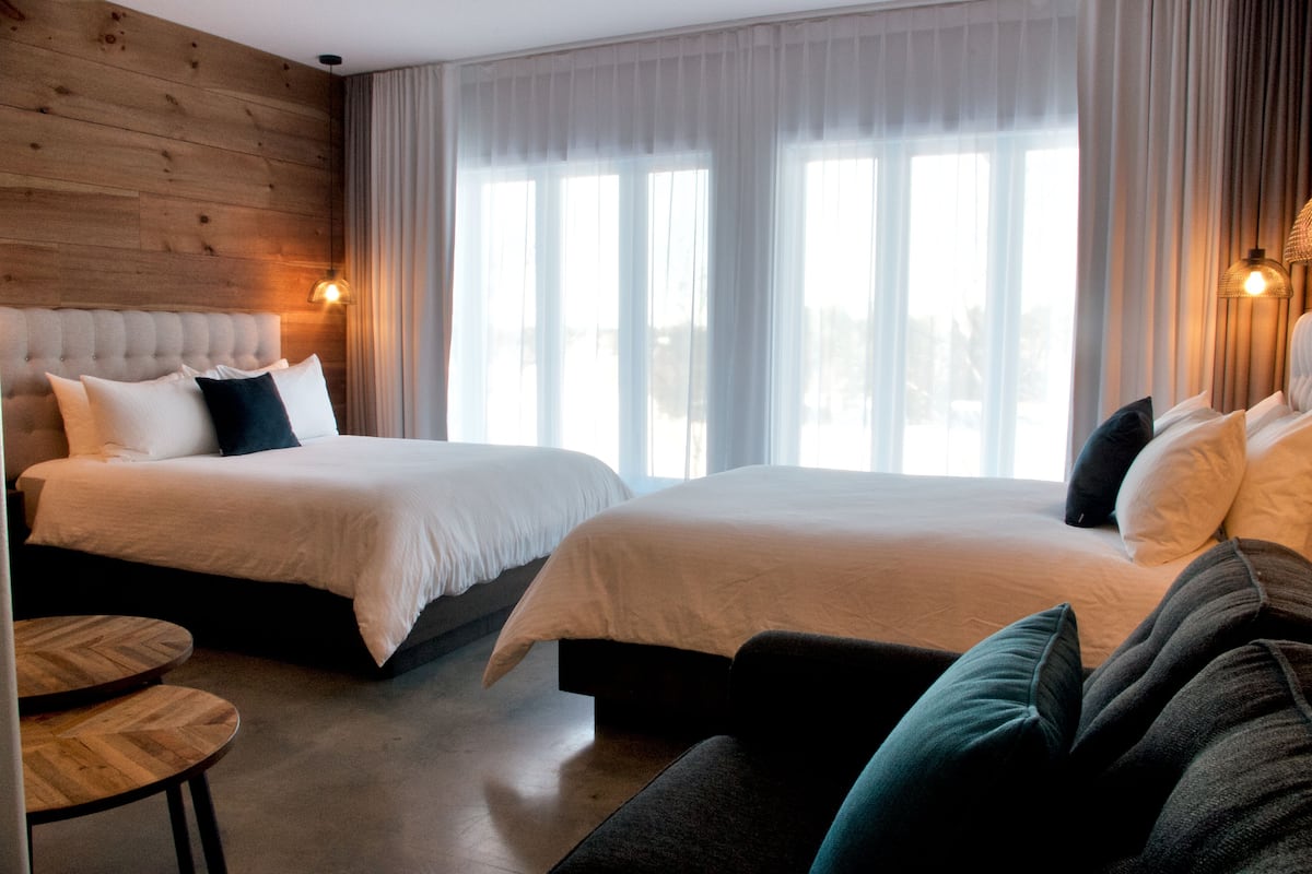 Habitación de hotel con dos camas, cabecera de madera, una mesa redonda de madera y vistas al exterior a través de amplias ventanas.