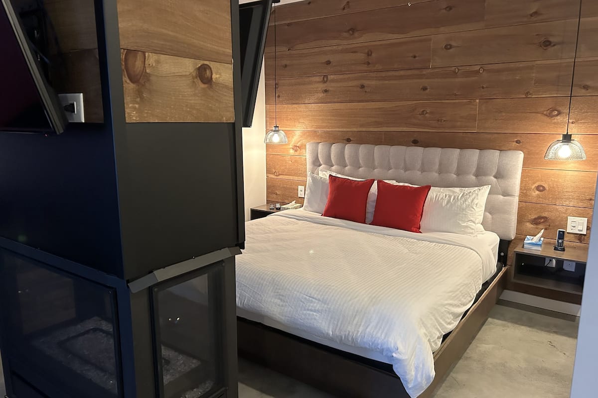 Un dormitorio moderno con cabecera de madera, cama con ropa blanca y almohadas rojas, mesita de noche con lámpara y televisión integrada.