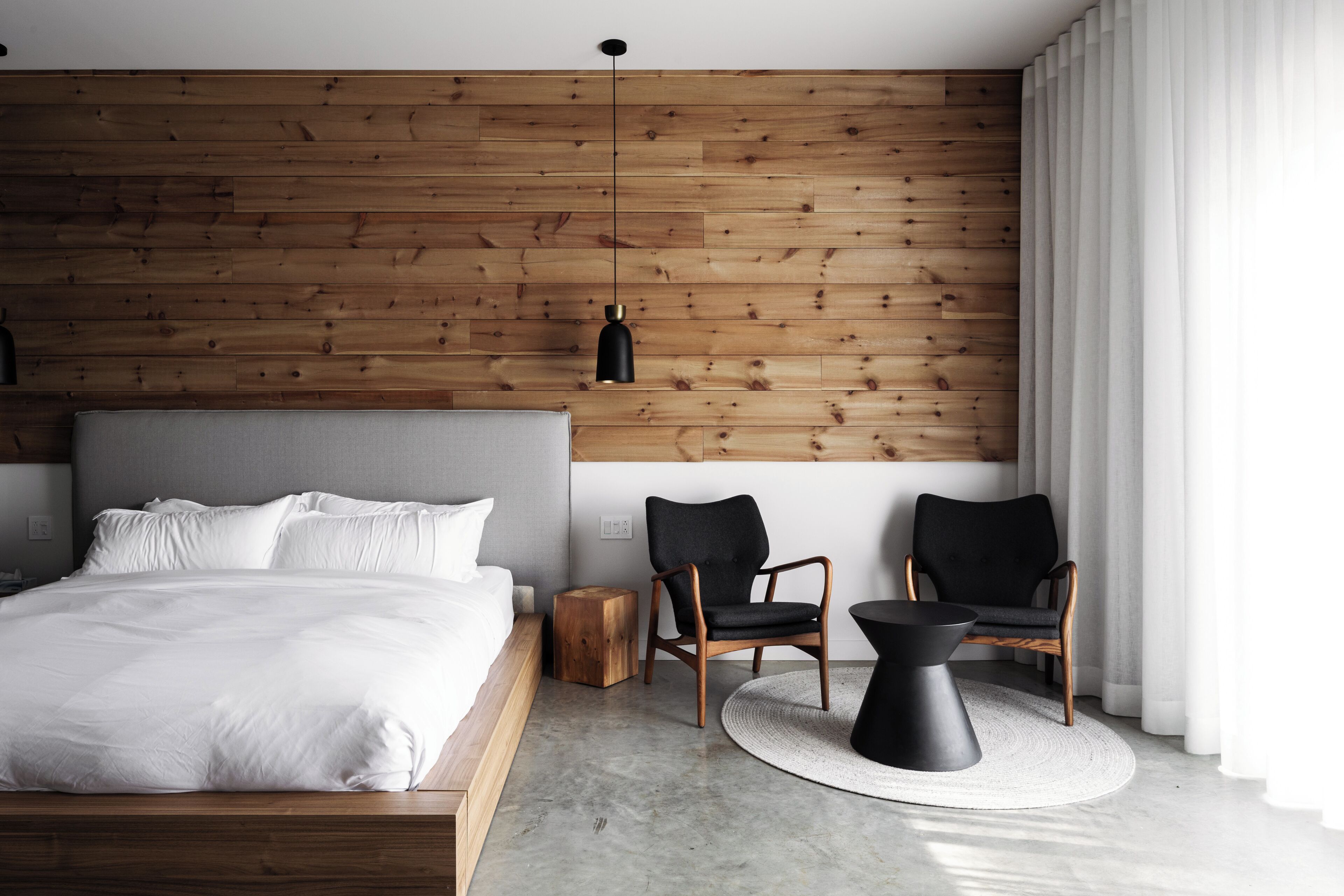 A modern bedroom with a wooden panel wall, a large bed, two chairs, a small side table, and a round coffee table.