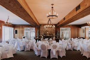 Un salón de banquetes con mesas redondas preparadas para una recepción de boda, con techo de madera y chimenea de piedra.