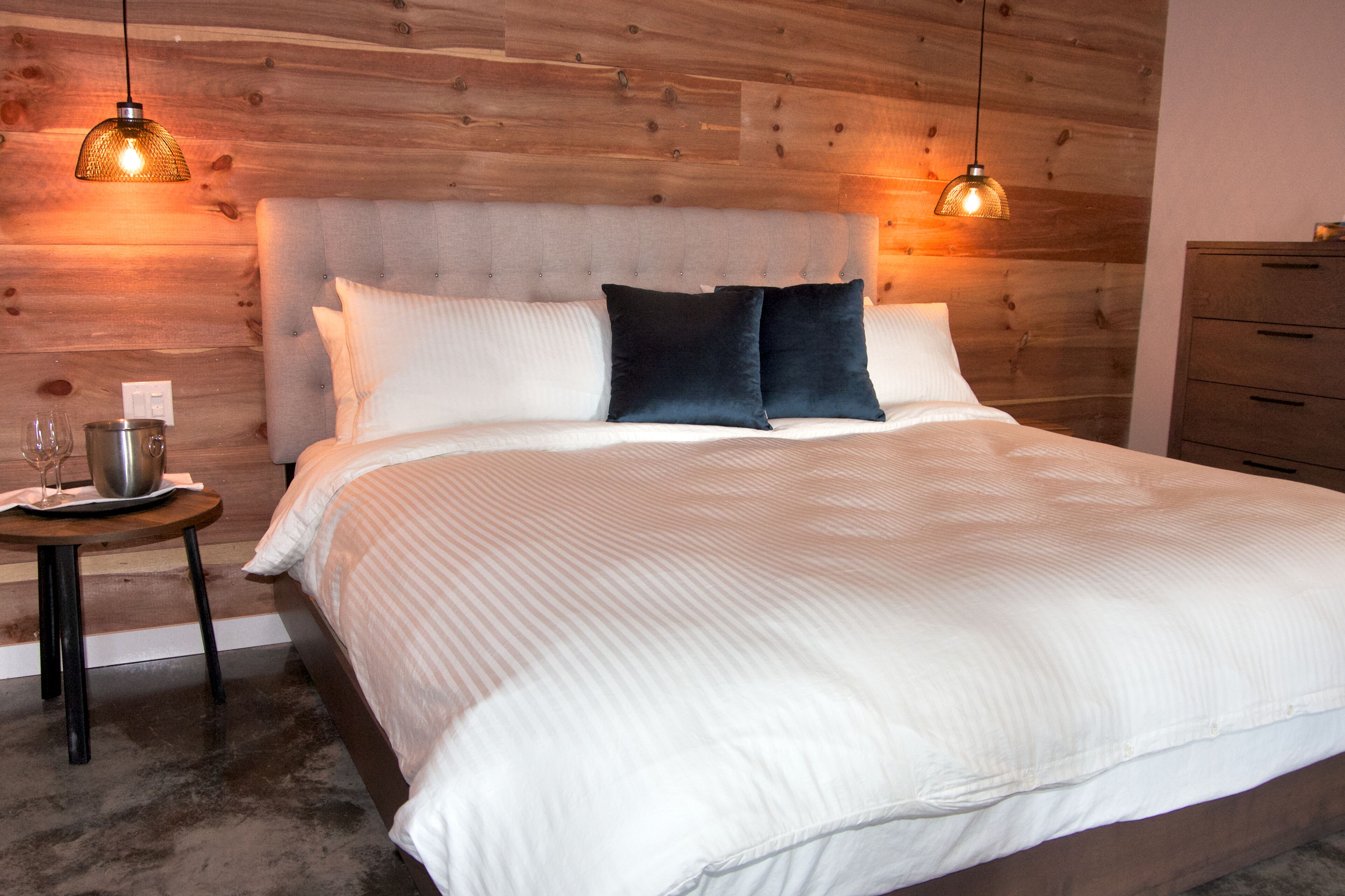 A neatly made bed with white and dark pillows, a wooden headboard, and two pendant lights.