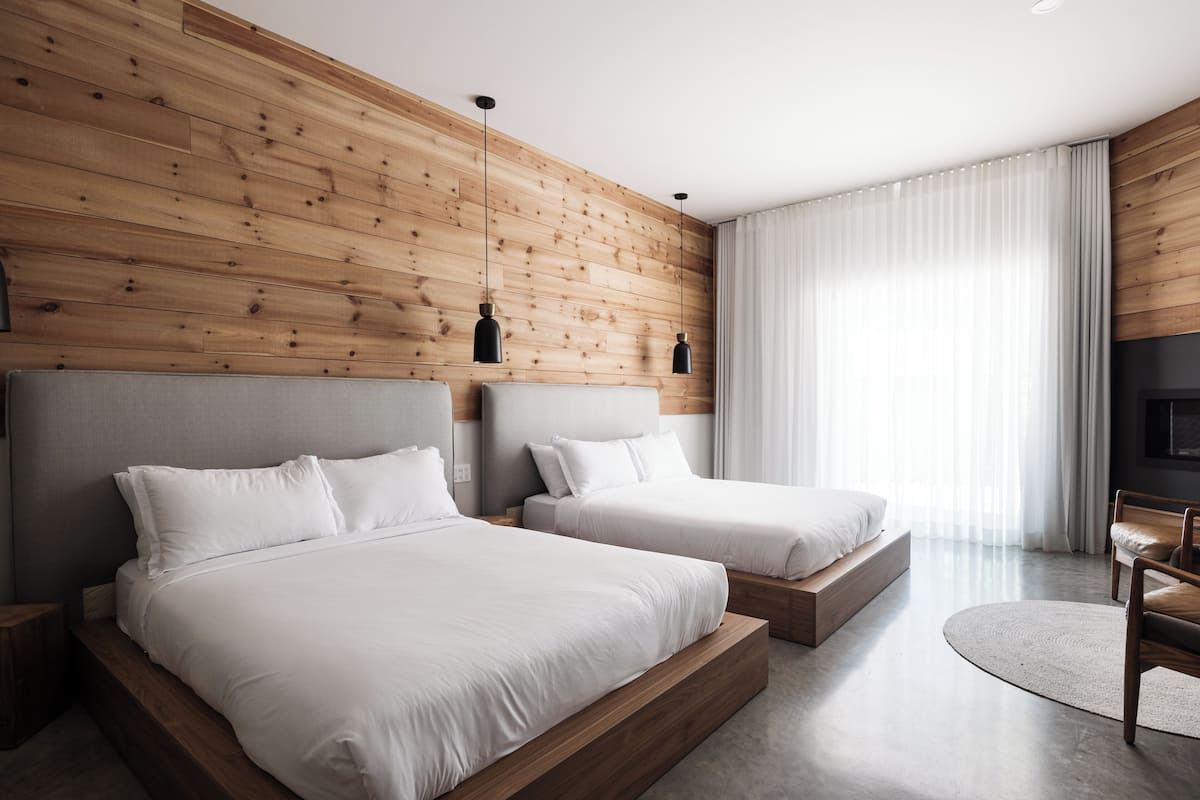 Habitación de hotel moderna con dos camas, paneles de madera, un ventanal con cortinas y un televisor de pantalla plana.
