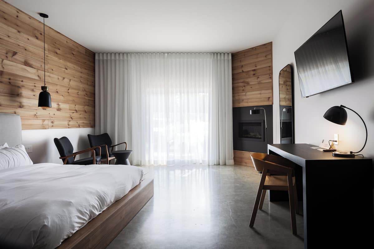 Una habitación de hotel moderna con pared de paneles de madera, una cama grande, un escritorio con una silla, un televisor de pantalla plana y un amplio ventanal con cortinas de gasa.