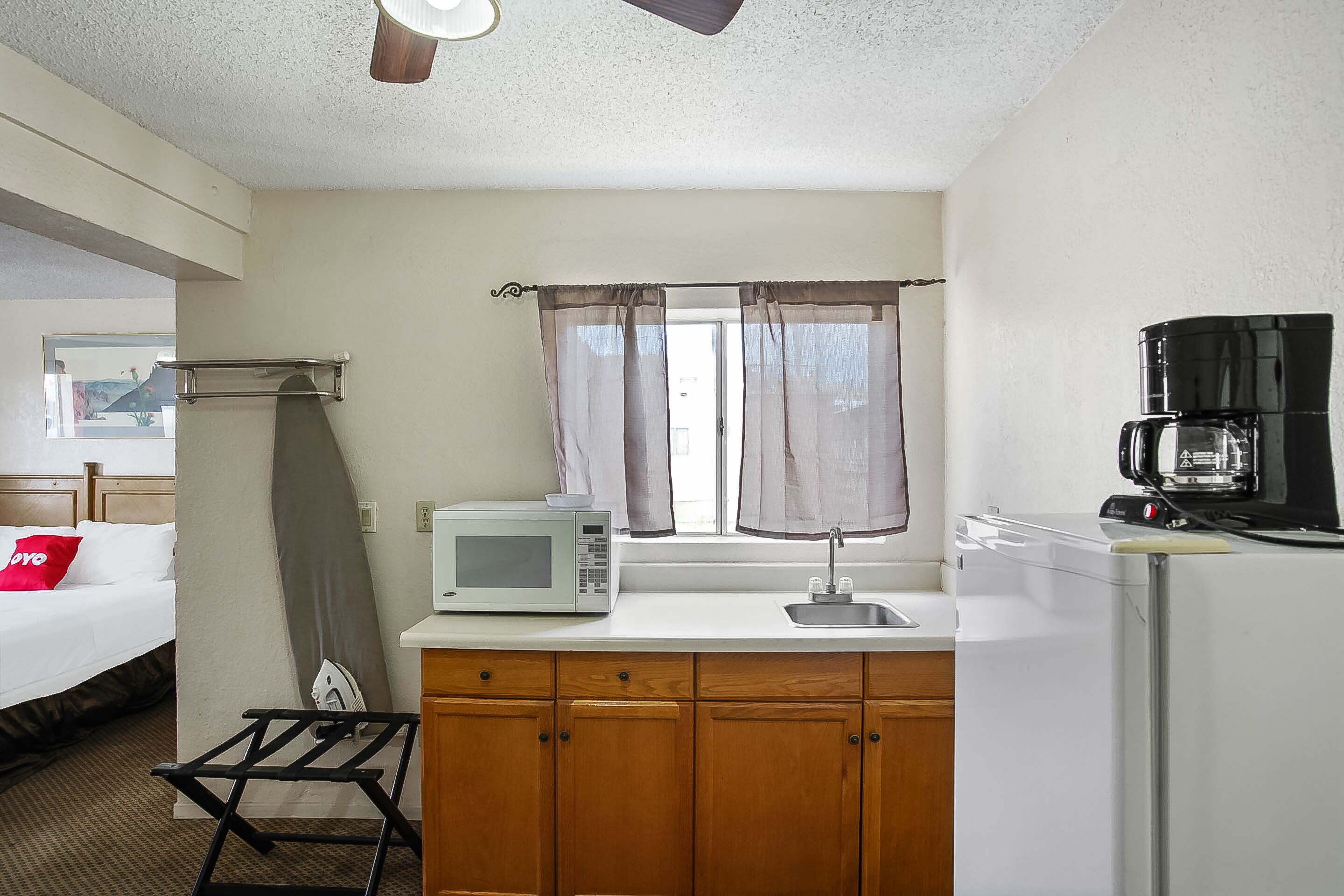 Habitación de hotel pequeña con kitchenette, una cama con almohada roja, microondas, cafetera y ventana con cortinas.