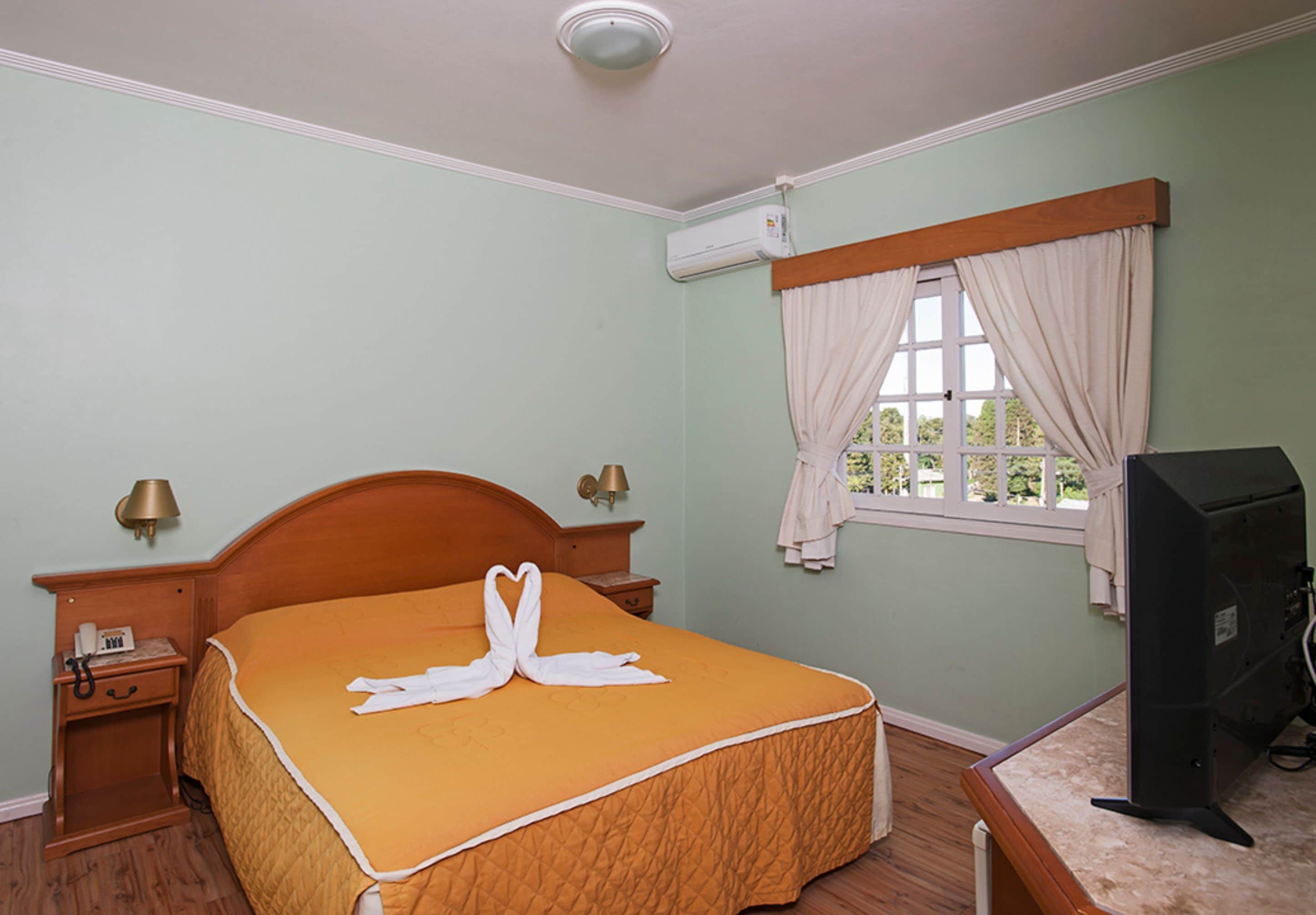 Una habitación de hotel con cama, lámparas de noche, televisor y ventana con cortinas.