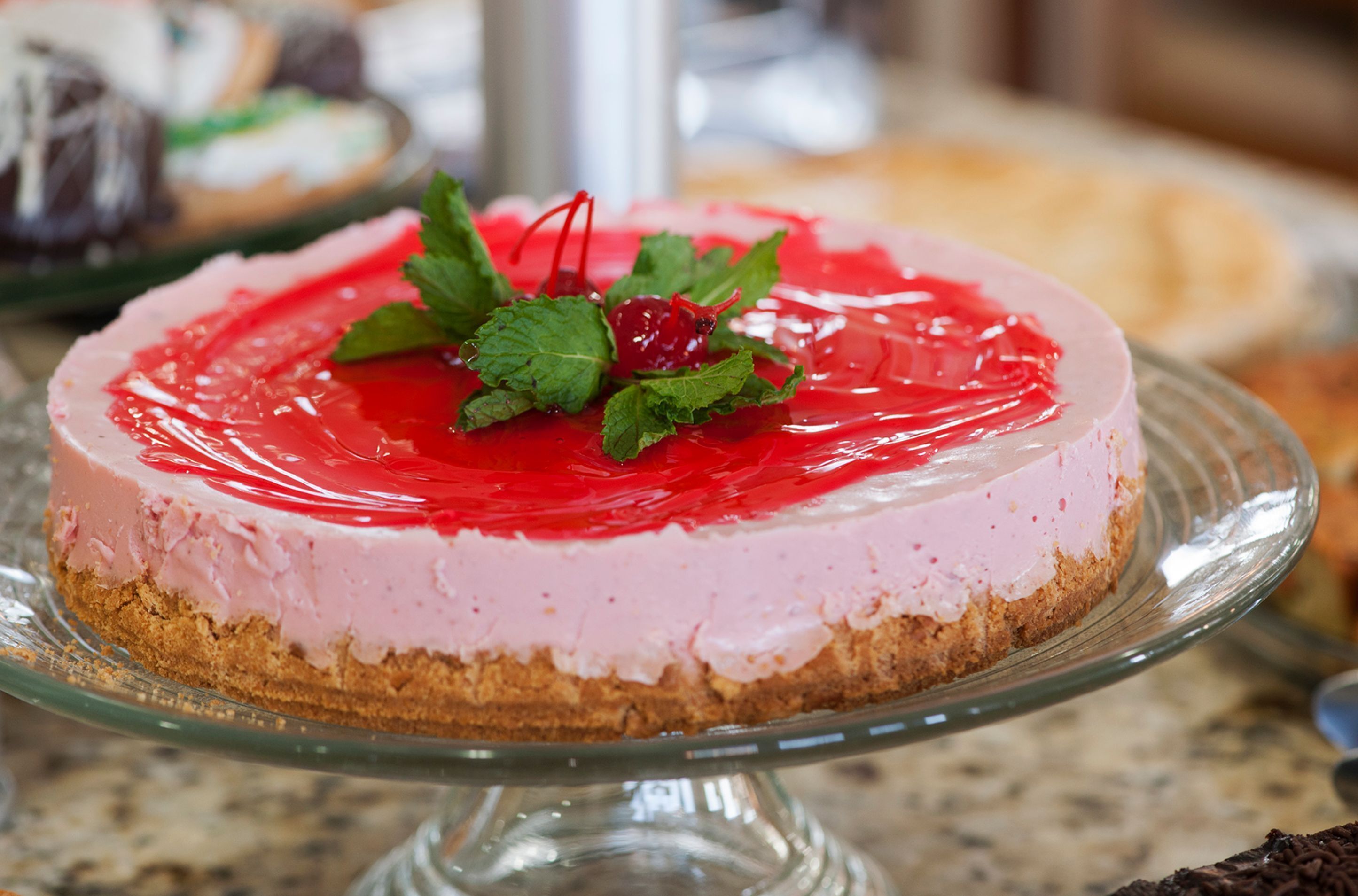 Un pastel de queso redondo con glaseado rojo y una cereza encima, servido sobre un expositor de vidrio para pasteles.