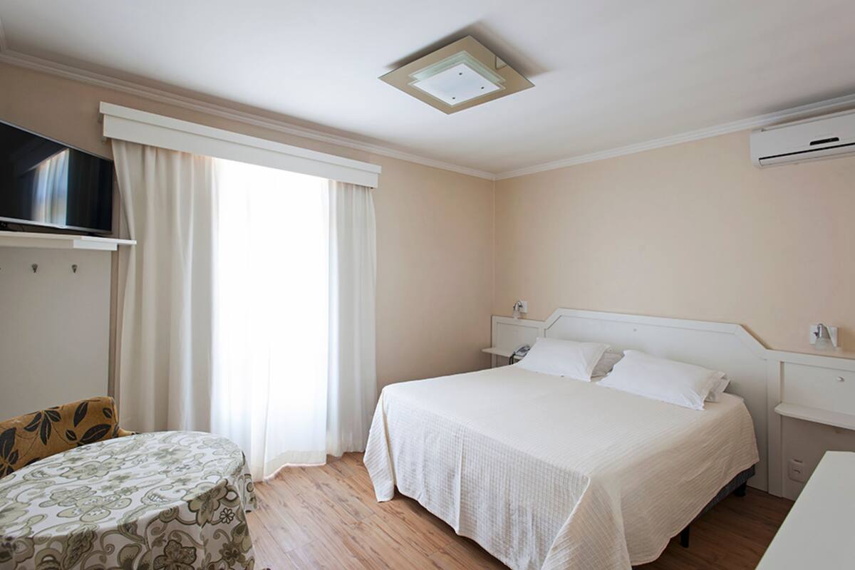 Ein Hotelzimmer mit einem Bett, einem Fernseher, einem kleinen Tisch und einem Fenster mit Vorhängen.