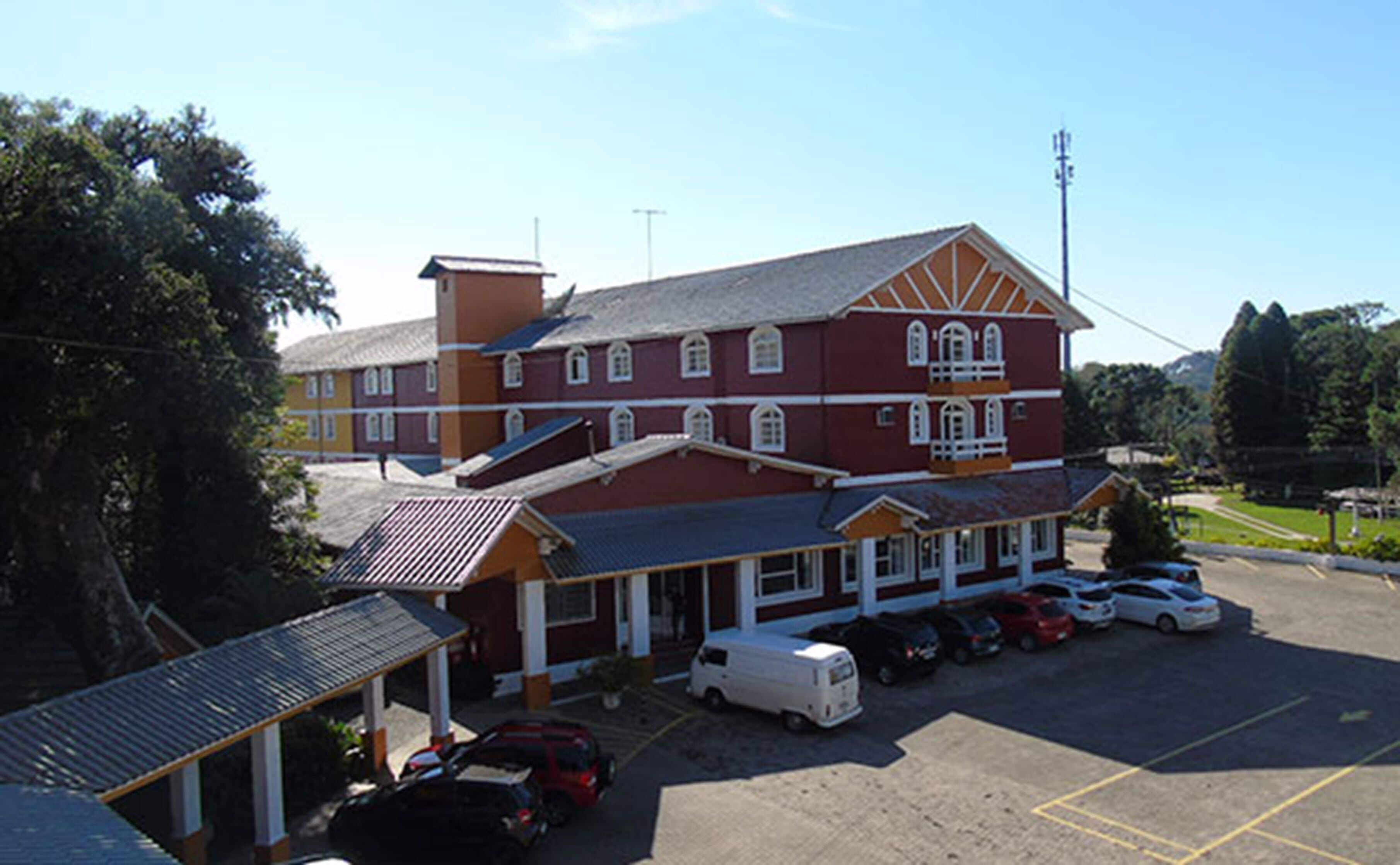 A large, multi-story building with a red facade and white trim, featuring a covered parking area with several cars and a white van.