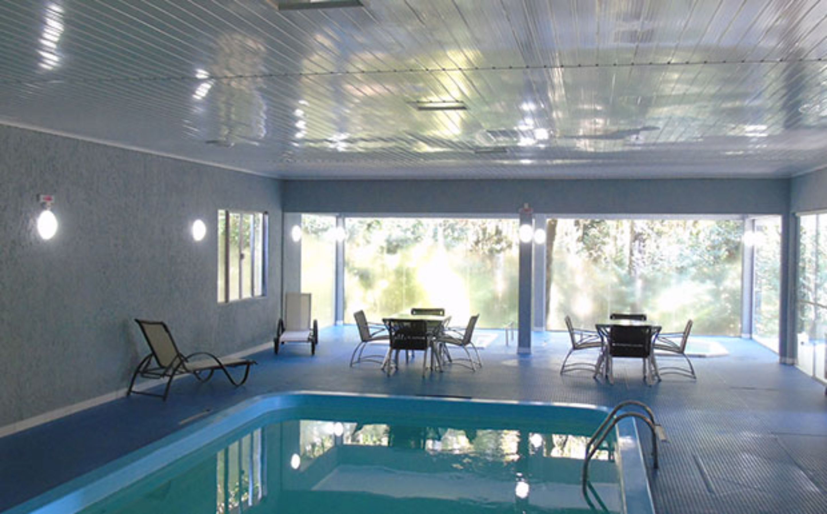 Piscina interna com vista clara para o exterior, cercada por espreguiçadeiras e mesas.