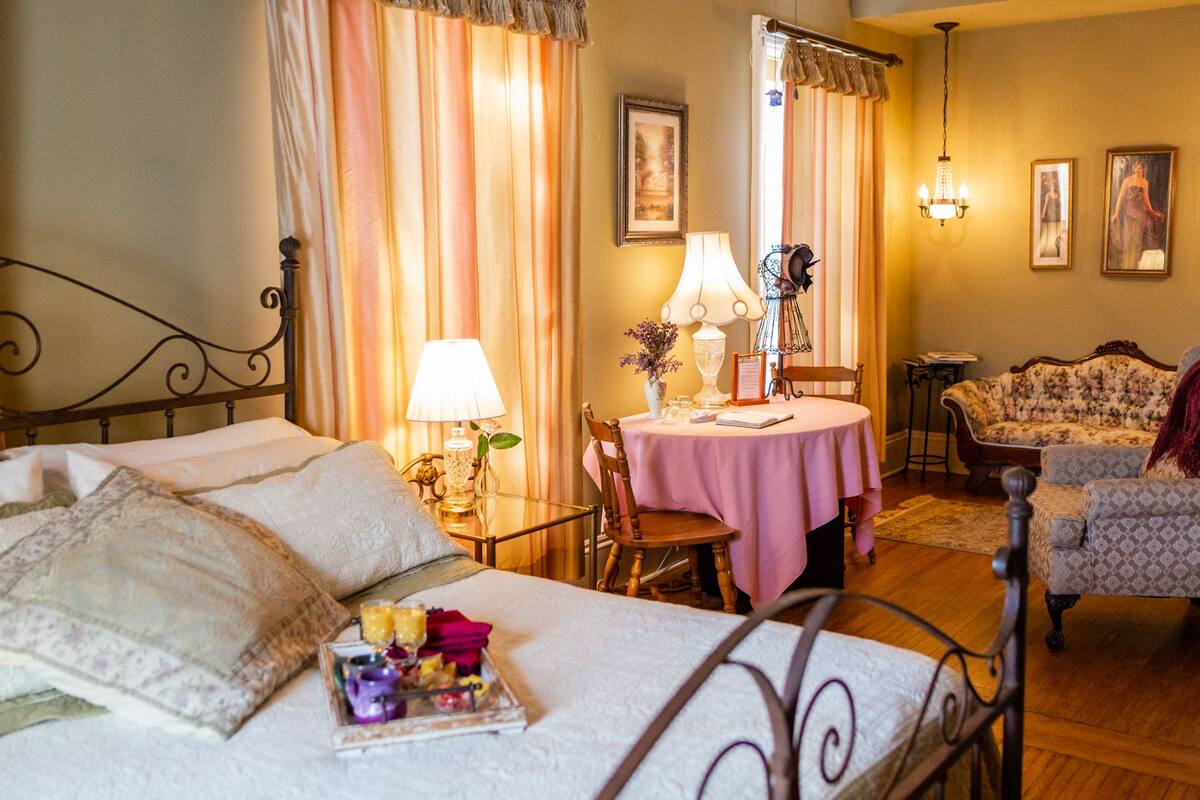 A hotel room with a bed, a dining table with a pink tablecloth, a chair, a sofa, and a small table with a lamp and flowers.