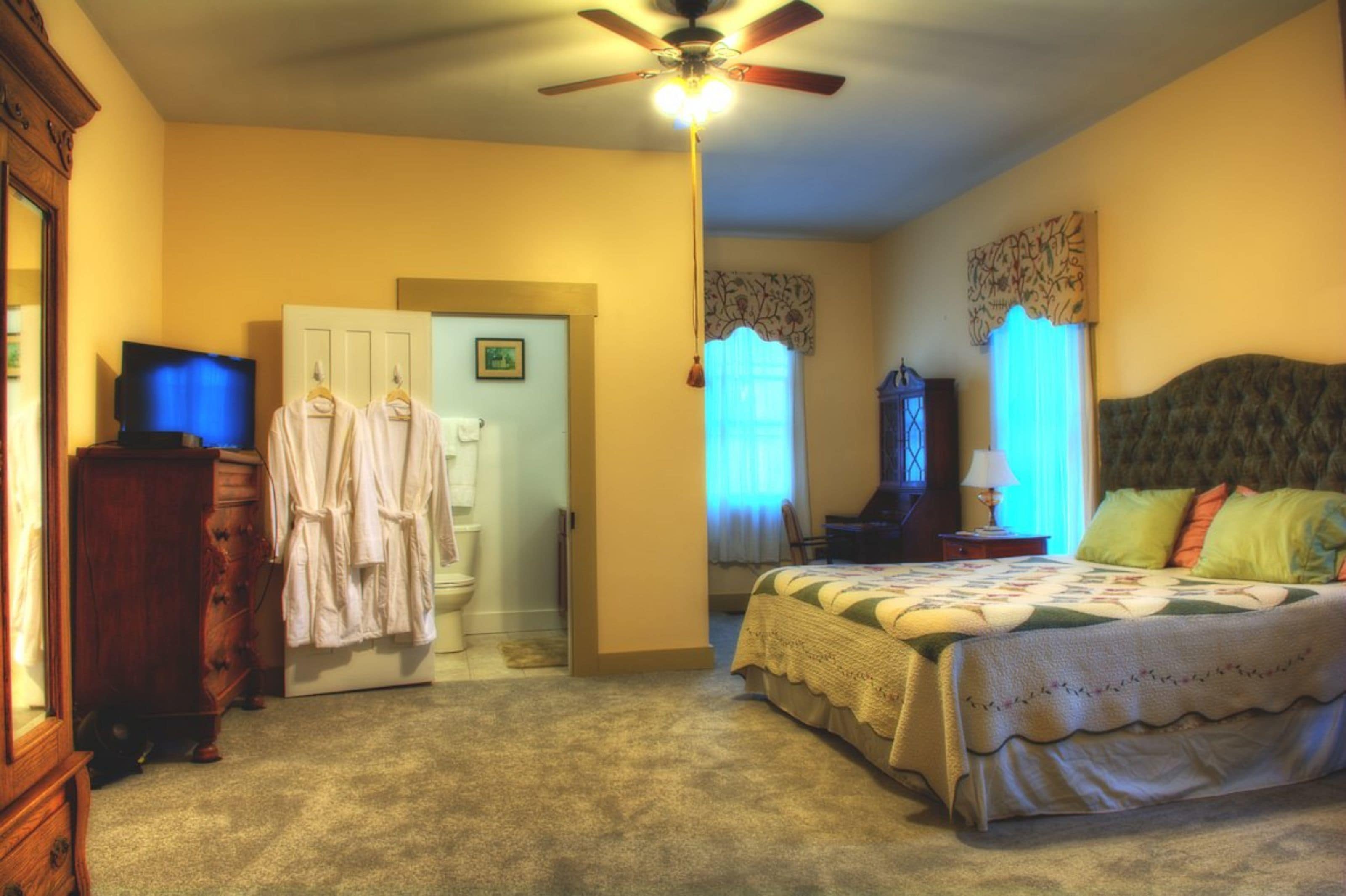 Un dormitorio con una cama, ventilador de techo, televisor, cómoda y una puerta que da a otra habitación.
