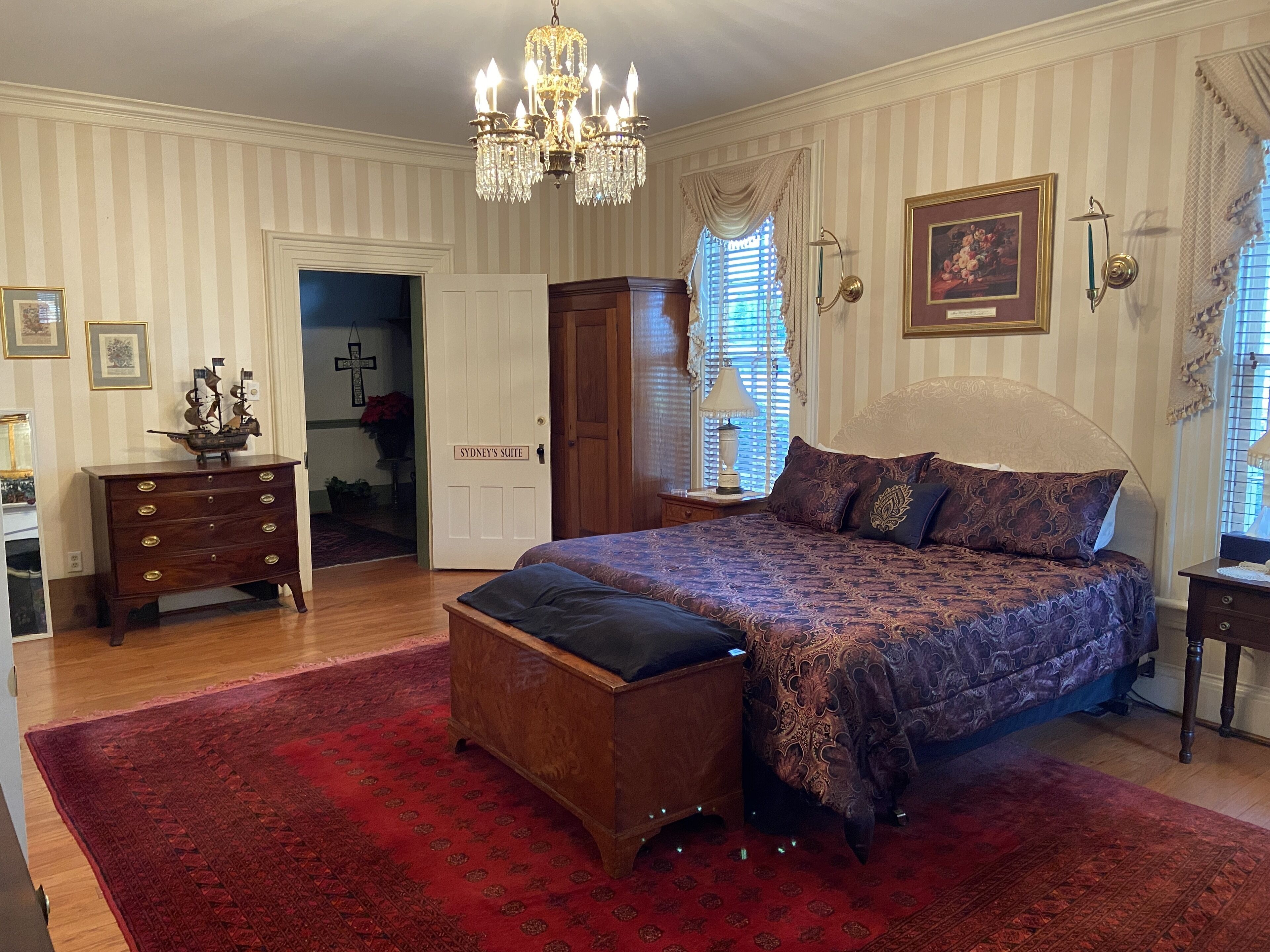 A bedroom with a large bed, a wooden dresser, a chandelier, and a door labeled "Sydney's Suite".
