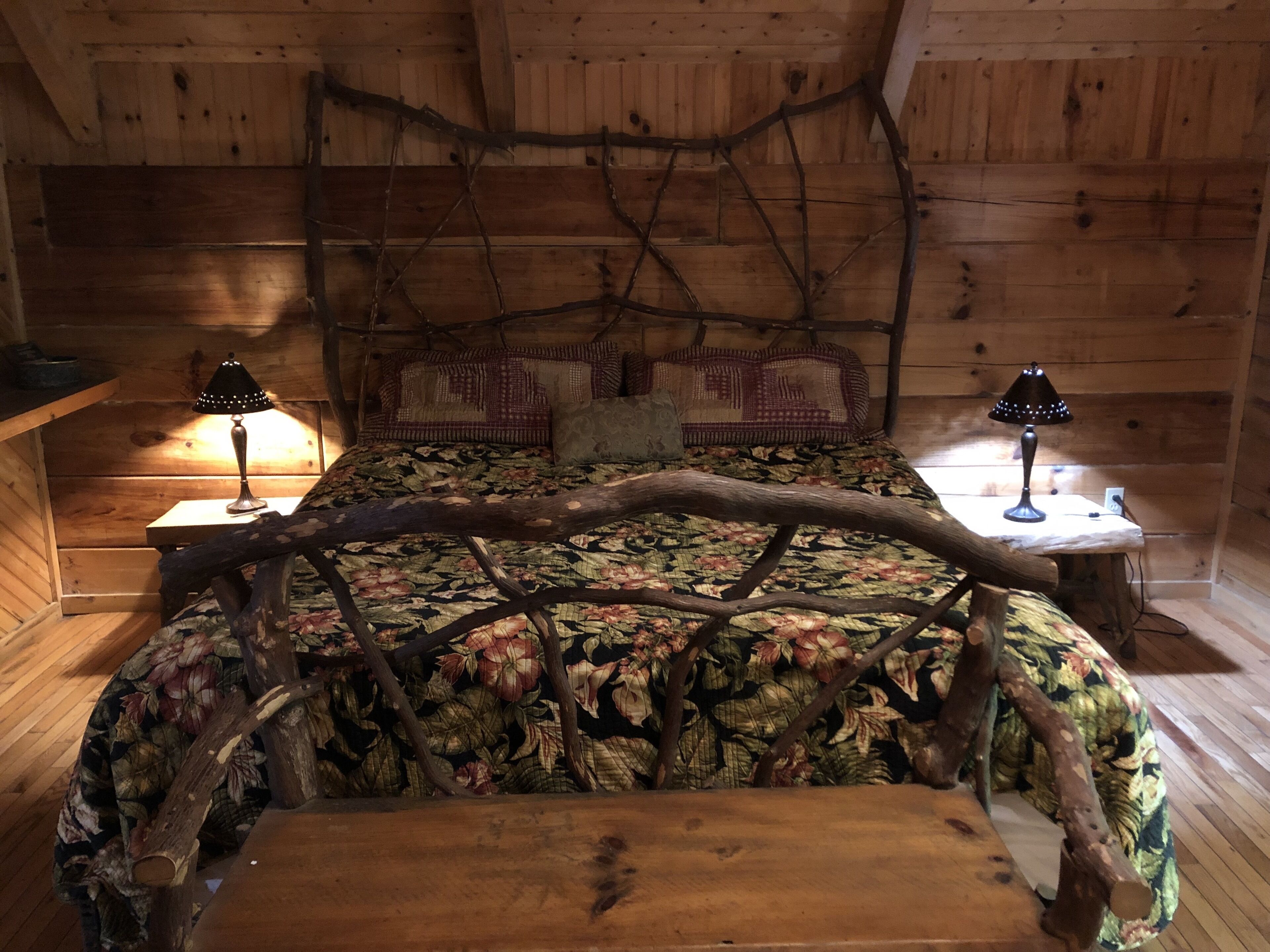 Ein rustikaler Holzraum mit einem Bett, das mit einem floralen Bettbezug versehen ist und einen großen Ast als Kopfteil hat. Auf Holzbeistelltischen stehen je eine Nachttischlampe.