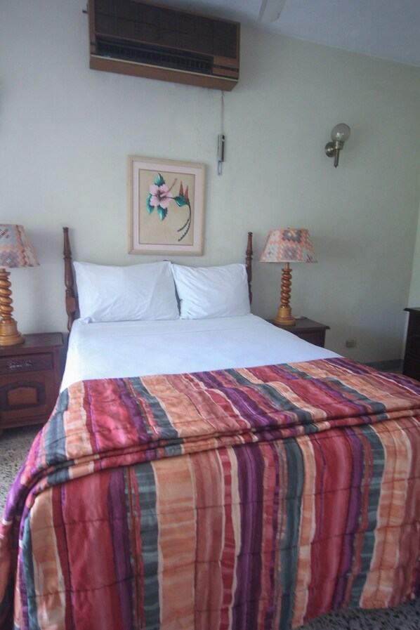 A neatly made bed with a colorful striped bedspread, two bedside tables with lamps, a wall-mounted air conditioner, and a framed picture above the bed. - Sahara Hostel (Montego Bay)