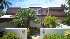 A two-story building with a brown tiled roof, a visible window, and a small garden area with palm trees and potted plants. - Burlingame Villa (Montego Bay)