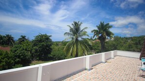 A rooftop terrace with a tiled floor, white railing, and a view of palm trees and greenery. - Burlingame Villa (Montego Bay)