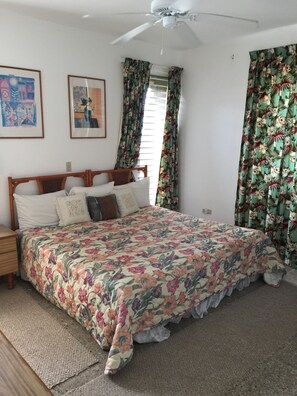 A bedroom with a wooden bed, floral bedspread, two framed artworks, a ceiling fan, and floral curtains.