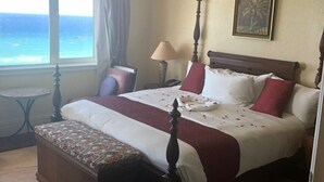 Une chambre à coucher comprenant un lit, une table de chevet, une lampe, un ventilateur de plafond, une fenêtre donnant sur l’océan et un tableau encadré accroché au mur.