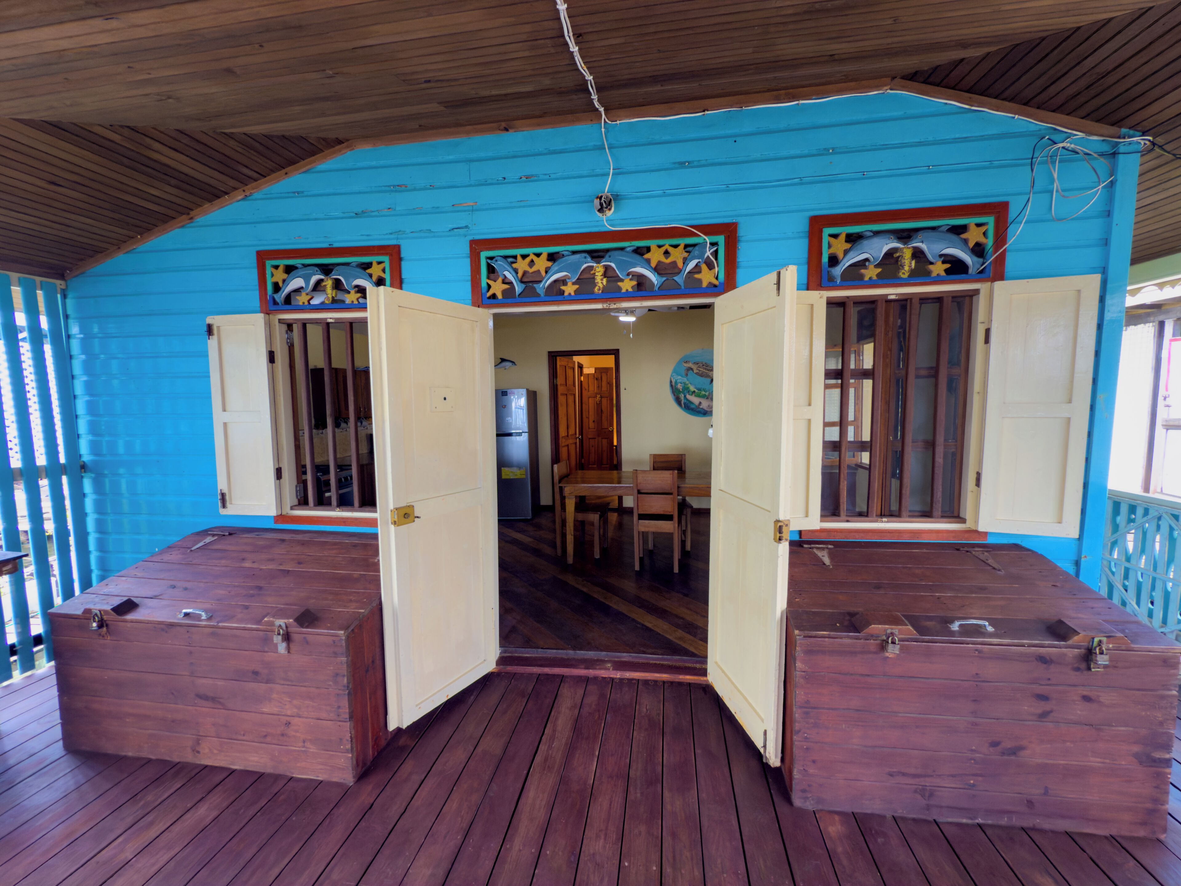 Une terrasse en bois, avec une façade bleue et des portes ouvertes donnant sur un espace intérieur comprenant une table à manger et des chaises.