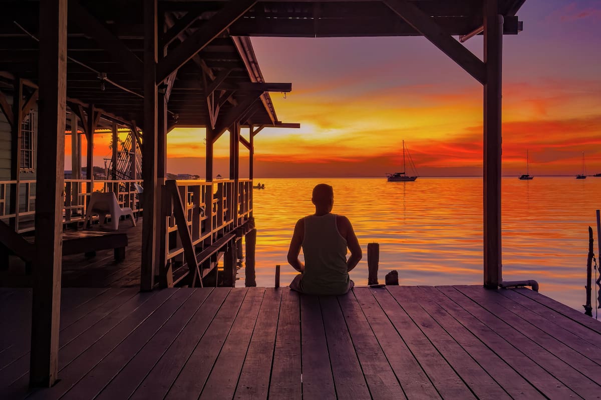 A wooden pier with a person sitting, overlooking a sunset over water with boats.