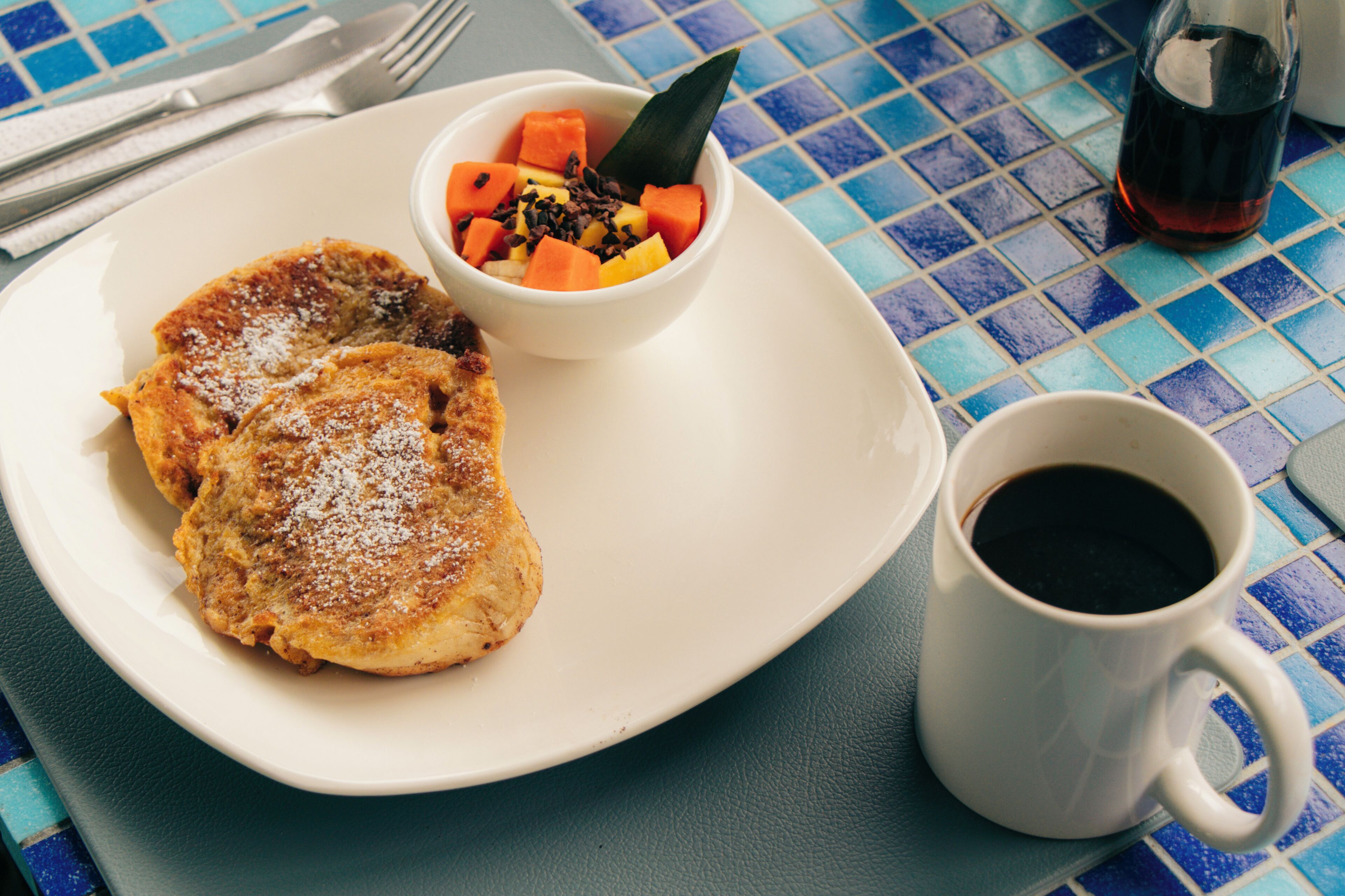A plate with a fried item and a bowl of fruit salad, a cup of coffee, and a glass of dark liquid on a tiled table.