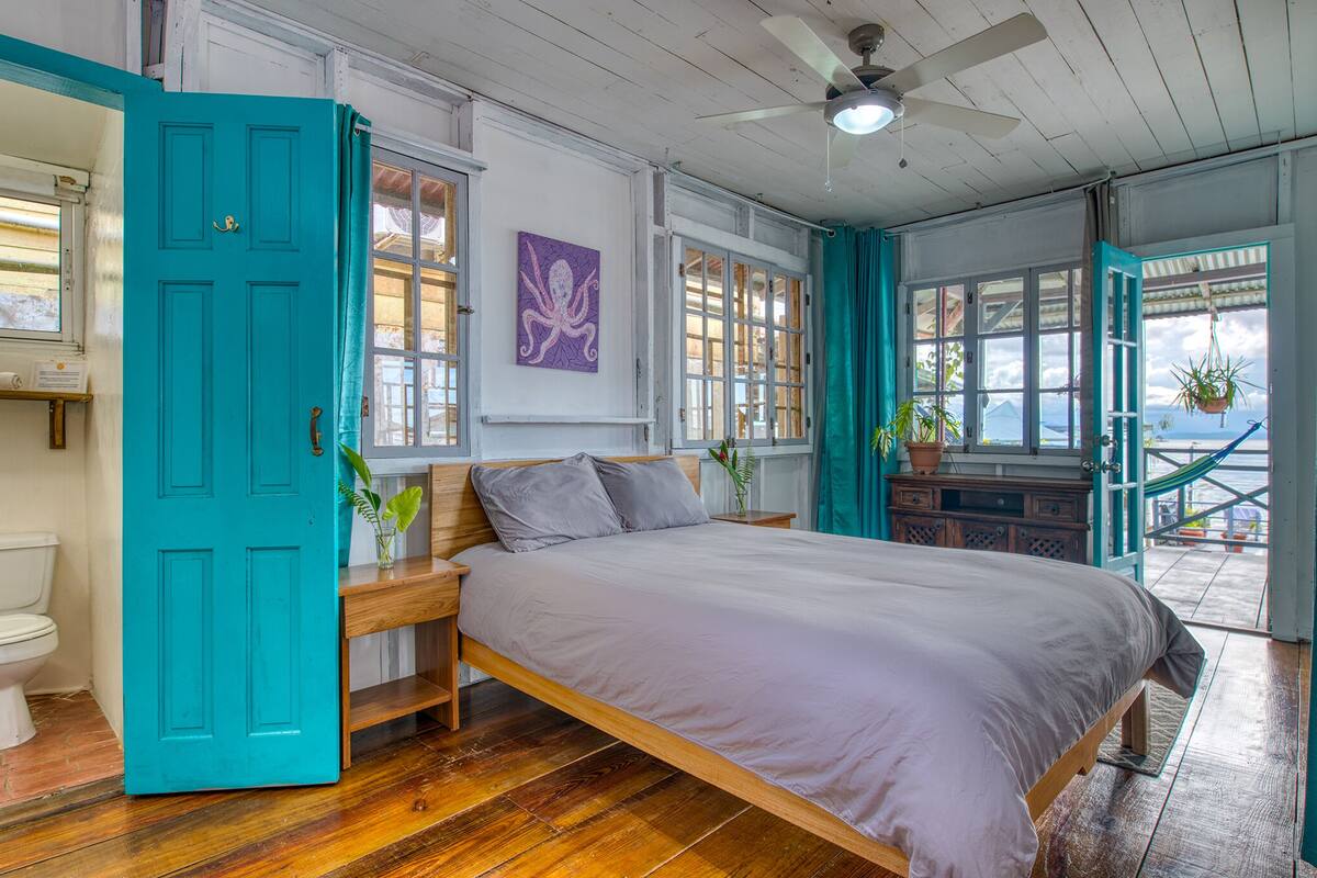 A room with a bed, a wooden nightstand, a teal door, and a view of the outside.
