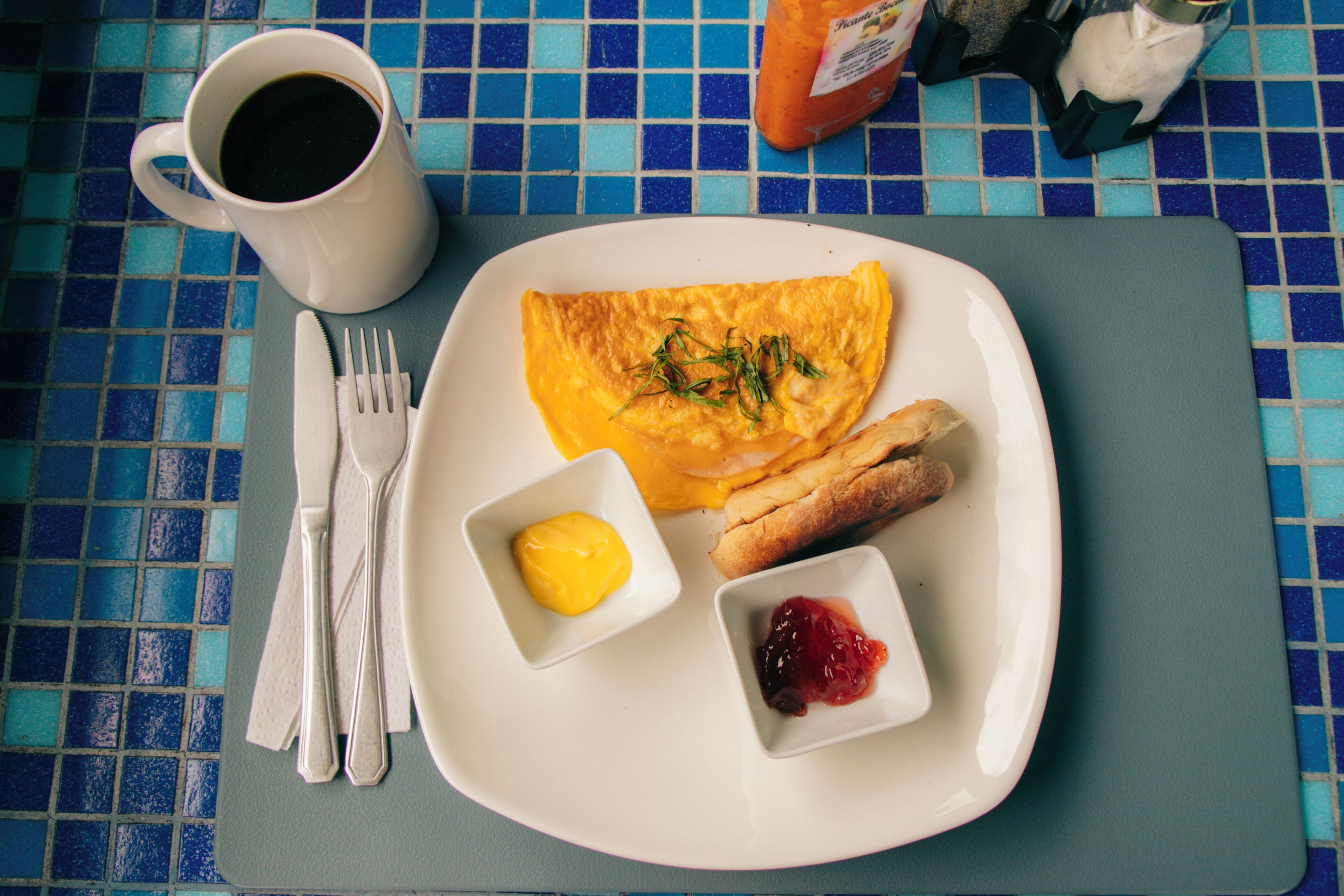 Une assiette contenant une omelette, des toasts, du beurre et de la confiture, une tasse de café et des couverts, le tout posé sur une table recouverte de carreaux bleus.