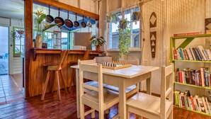 A cozy room with a wooden table and chairs, a bookshelf, and a window with hanging plants. - Saigon Bay Bed & Breakfast (Bocas del Toro)