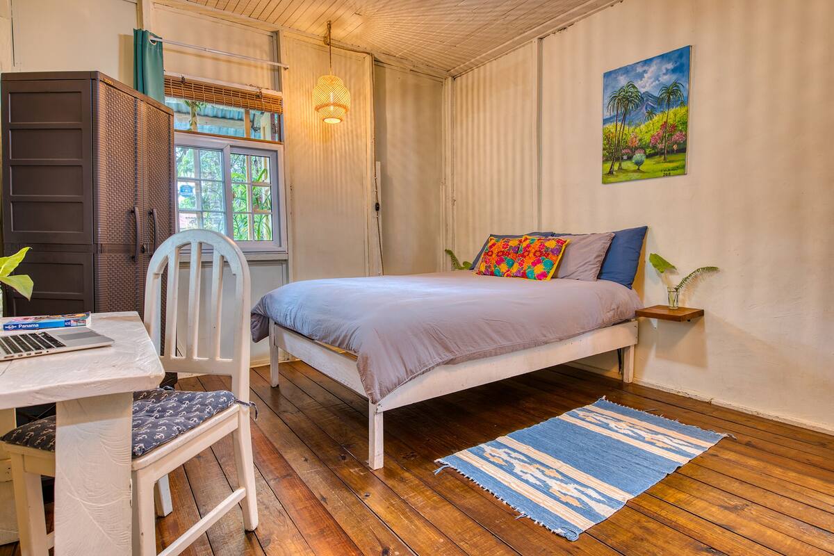 A bedroom with a bed, a desk with a laptop, a chair, a window, a painting, and a wooden floor.