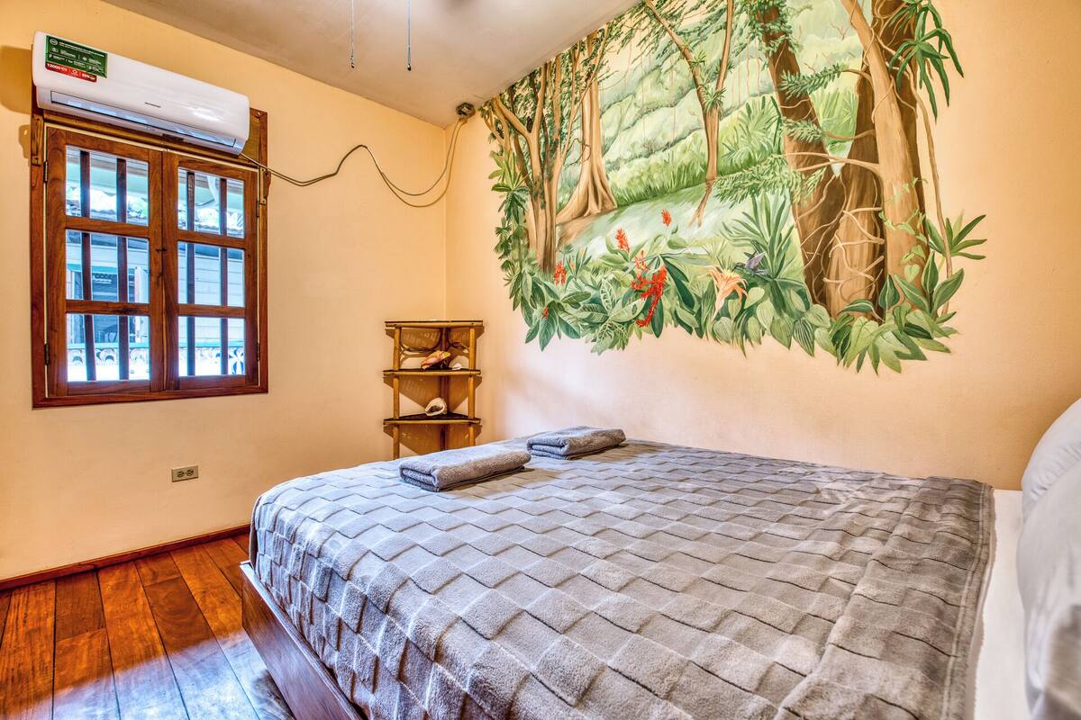 A bedroom with a bed, a window, an air conditioning unit, a shelf, and a mural of a forest scene.