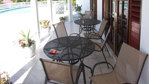 Eine überdachte Terrasse mit rundem Tisch und Stühlen, Blumentöpfen und Blick auf einen Pool und viel Grün.