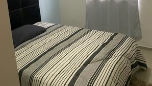 A bedroom with a bed, a headboard, a window with a sheer curtain, and a wall-mounted air conditioning unit. - Casa Elena (Mazatlán)