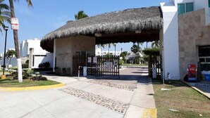 A building with a thatched roof entrance, palm trees, and a paved walkway leading to a gated area. - Casa Elena (Mazatlán)