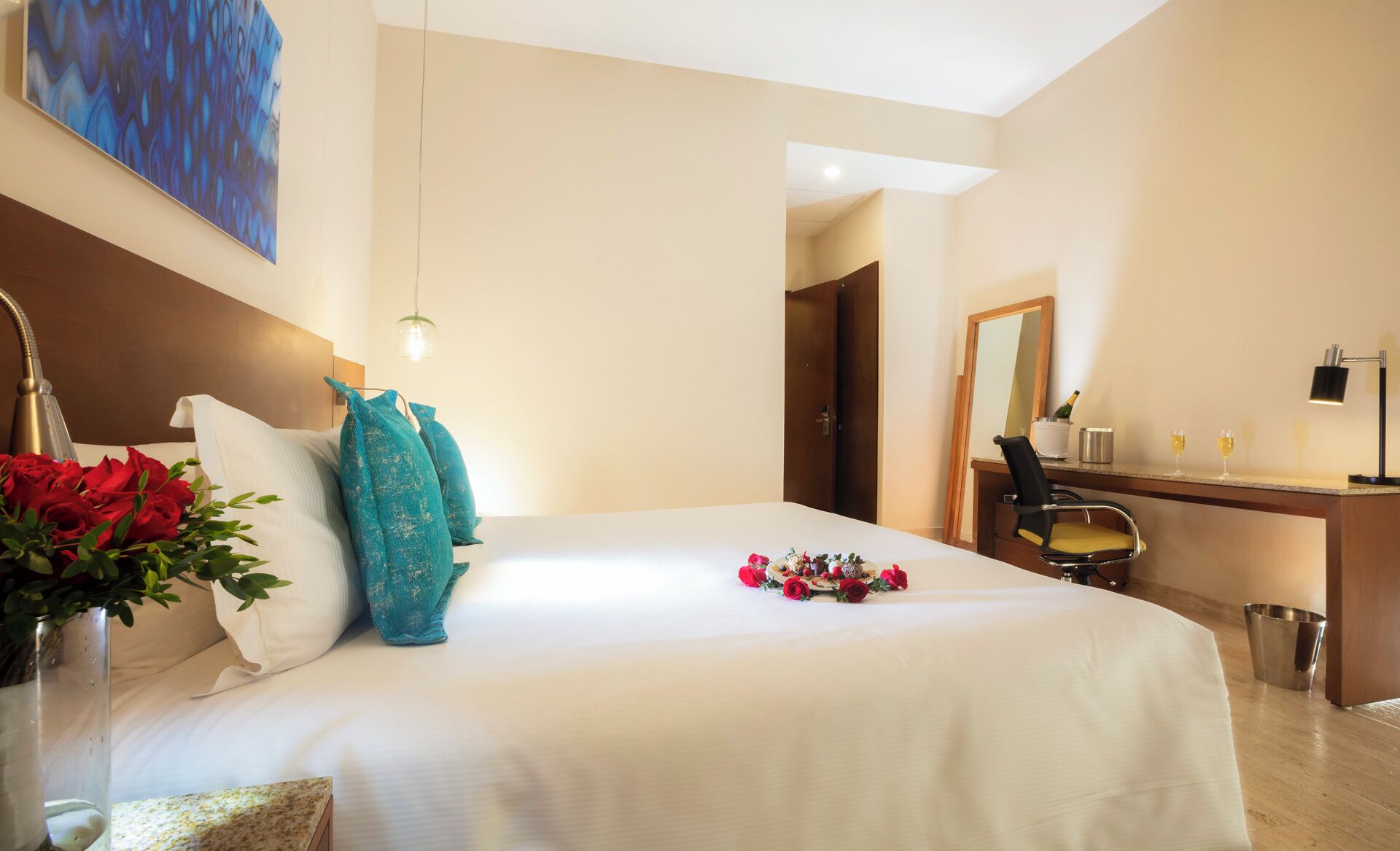 A hotel room with a bed, a desk, a mirror, and a vase with flowers.