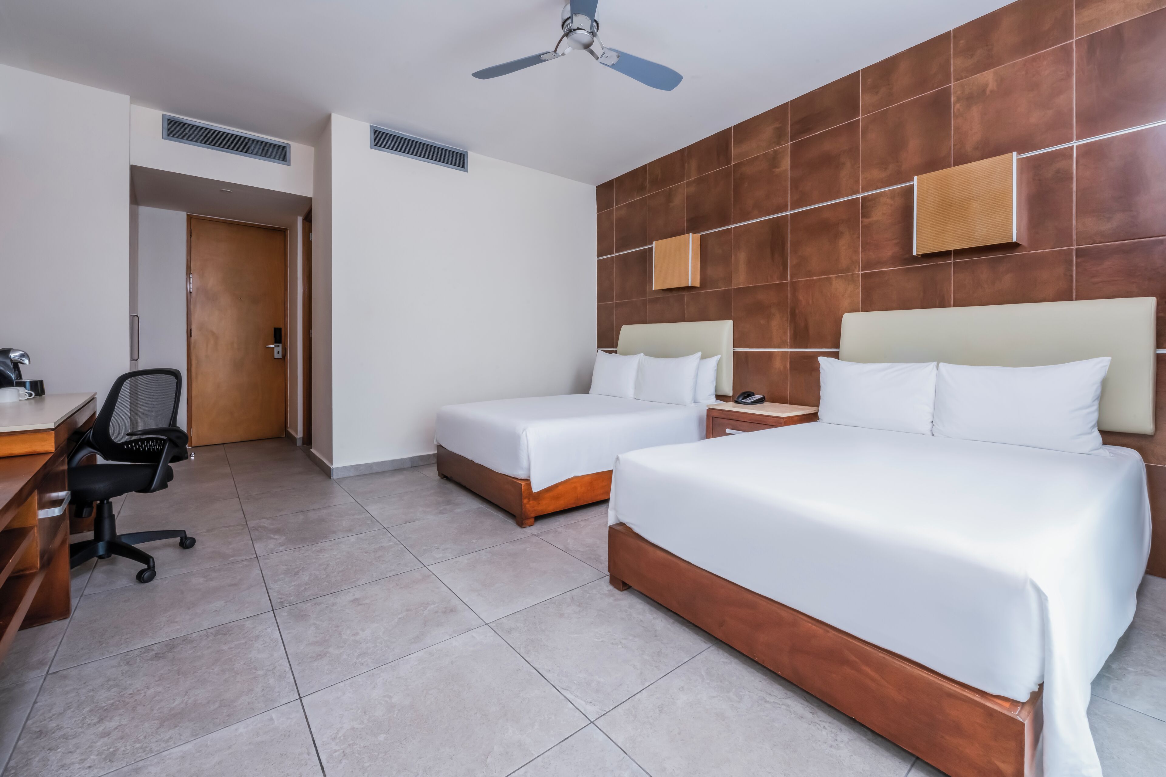 Una habitación de hotel con dos camas, un escritorio y una silla, y un ventilador de techo.