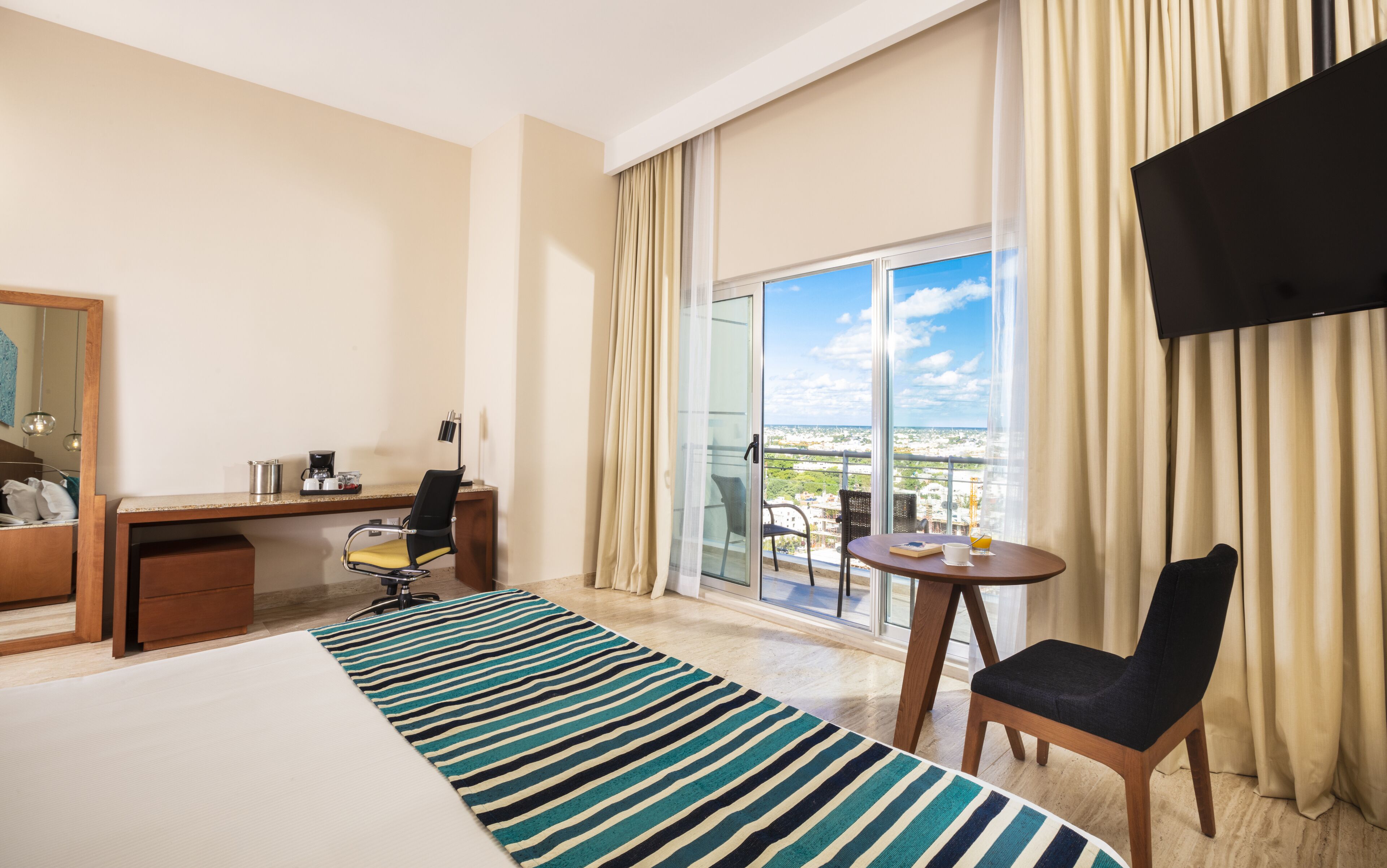 A hotel room with a bed, a desk with a chair, a small table with a chair, a TV, and a balcony with a view.