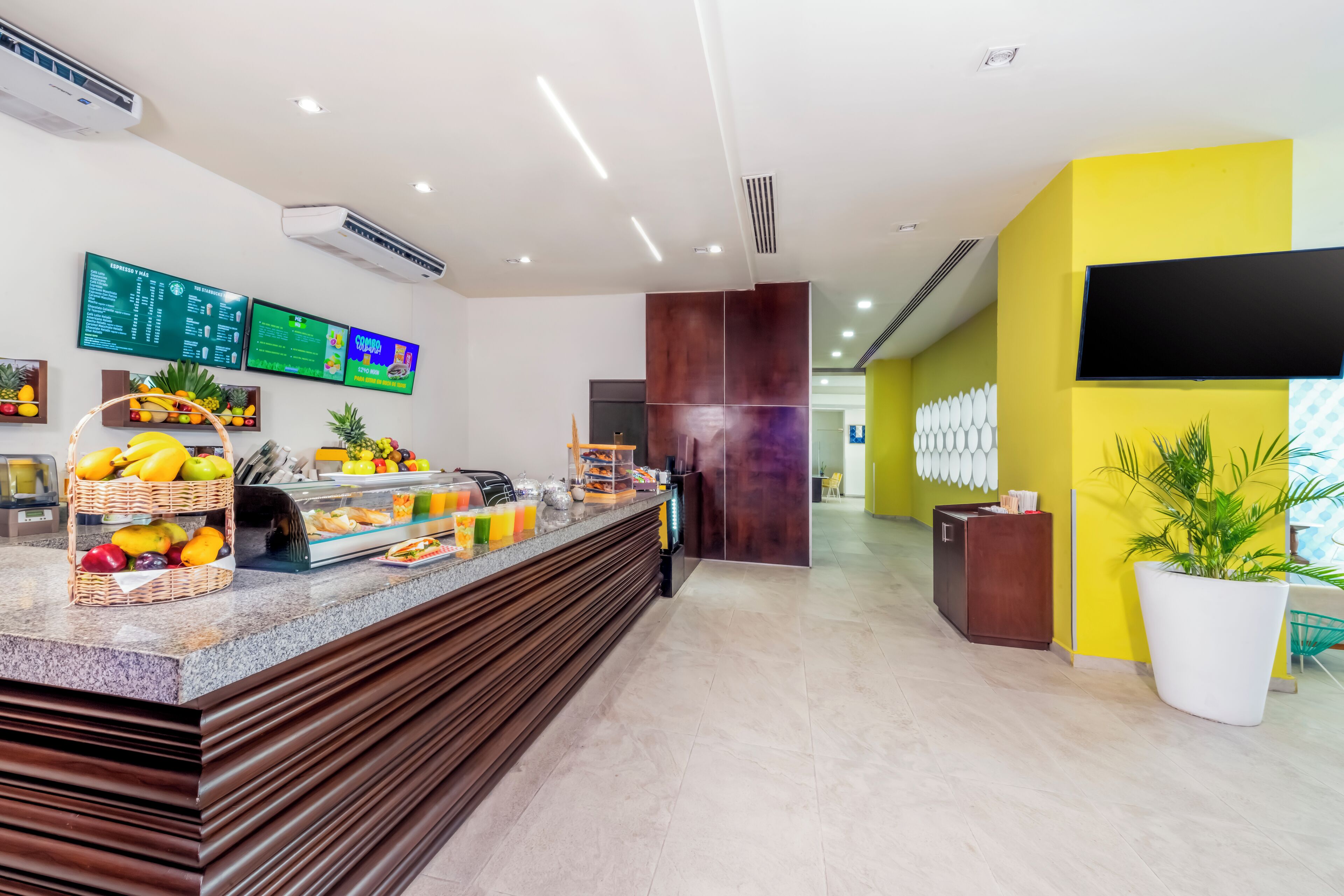 A modern hotel reception area with a fruit display, digital menu boards, and a bright yellow accent wall.