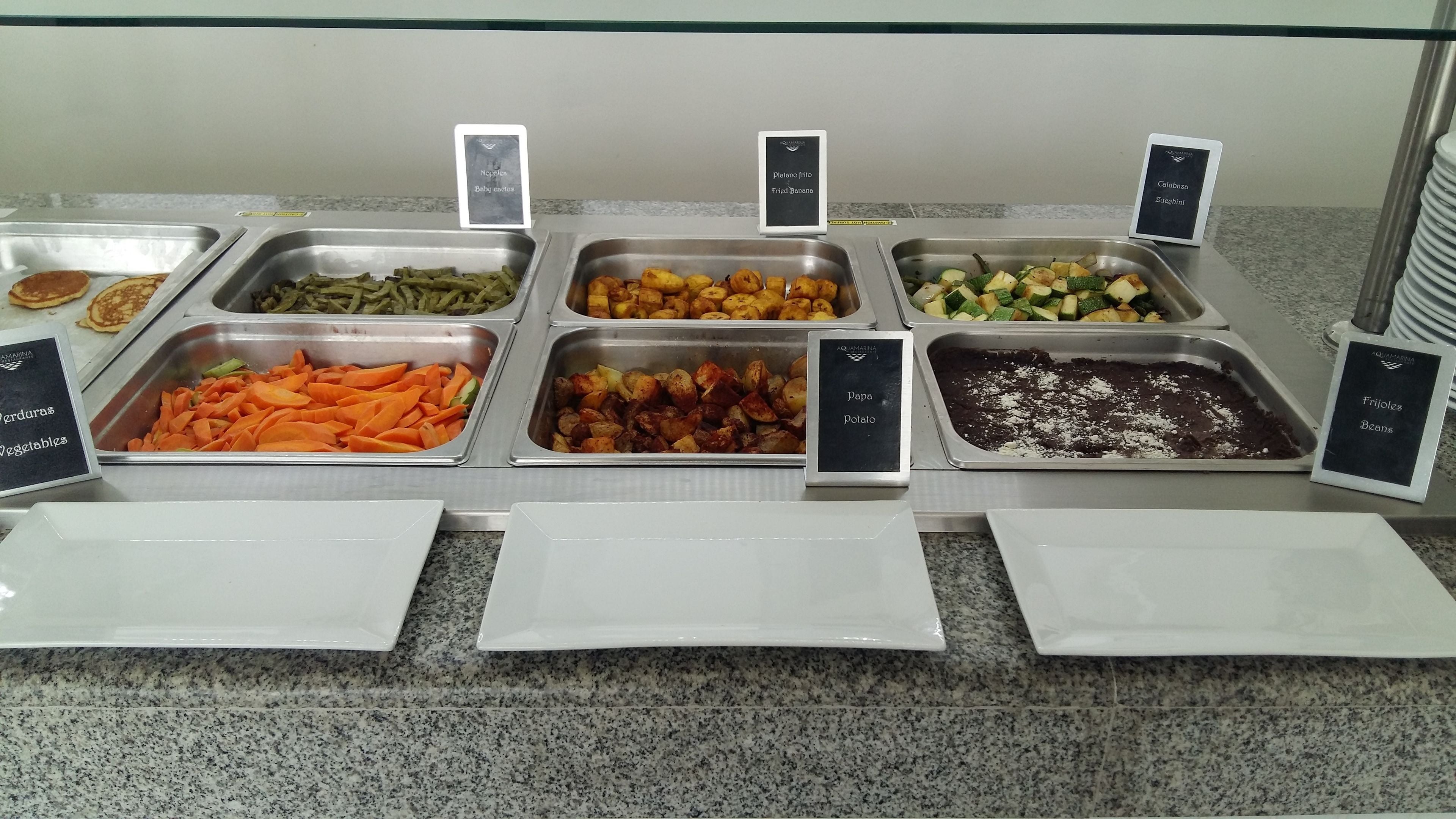 Buffet com diversas opções de comida, incluindo milho tenro, banana frita, abobrinha, cenoura, batatas e feijão.