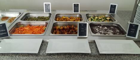 Buffet com diversas opções de comida, incluindo milho tenro, banana frita, abobrinha, cenoura, batatas e feijão.
