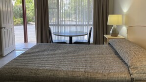 A hotel room with a bed, a table with chairs, a lamp, and a view of greenery through the window.
