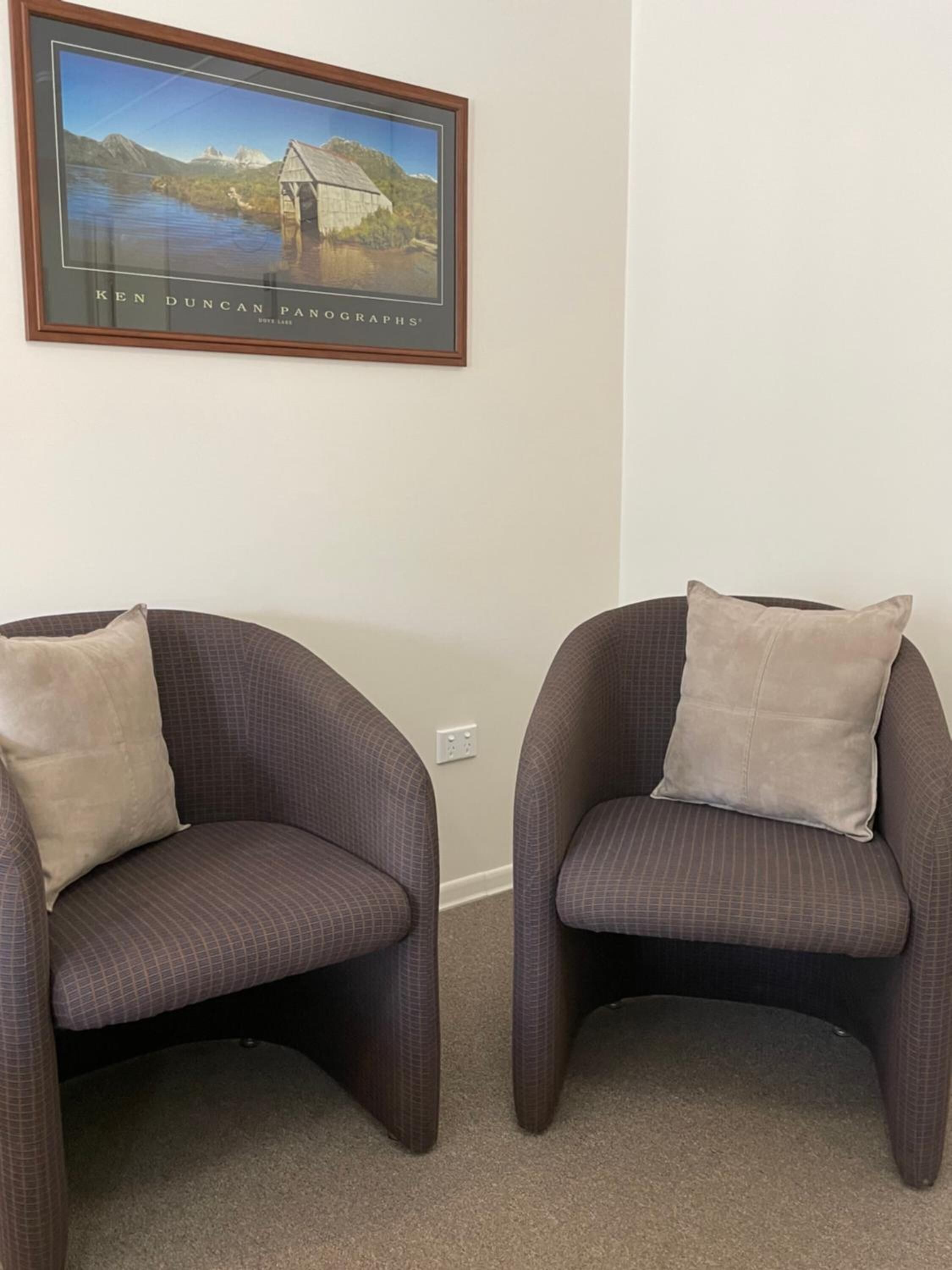 Two armchairs with cushions, a framed picture of a bridge, and a plain wall.